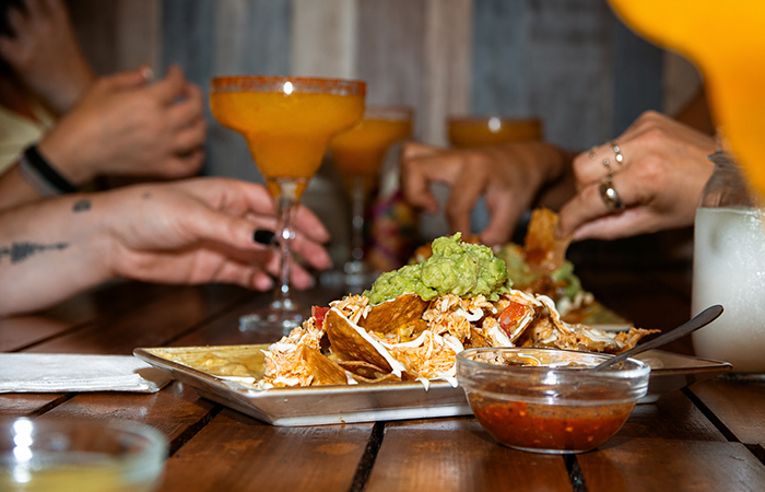 Nachos mexicanos para compartir Nachos mexicanos con guacamole y salsa servidos en una mesa junto a bebidas y comensales.